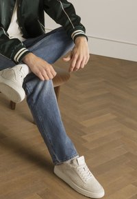 Weiße Wildledersneaker mit strukturierter Oberfläche, kontrastierenden Schnürsenkeln, runder Zehenpartie, getragen mit hellblauen Jeans und einer grünen Bomberjacke.