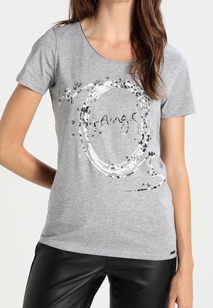 Kobieta w szarym t-shircie z krótkim rękawem z srebrnym cekinowym kołem i napisem "strange", w połączeniu z czarnymi skórzanymi spodniami.