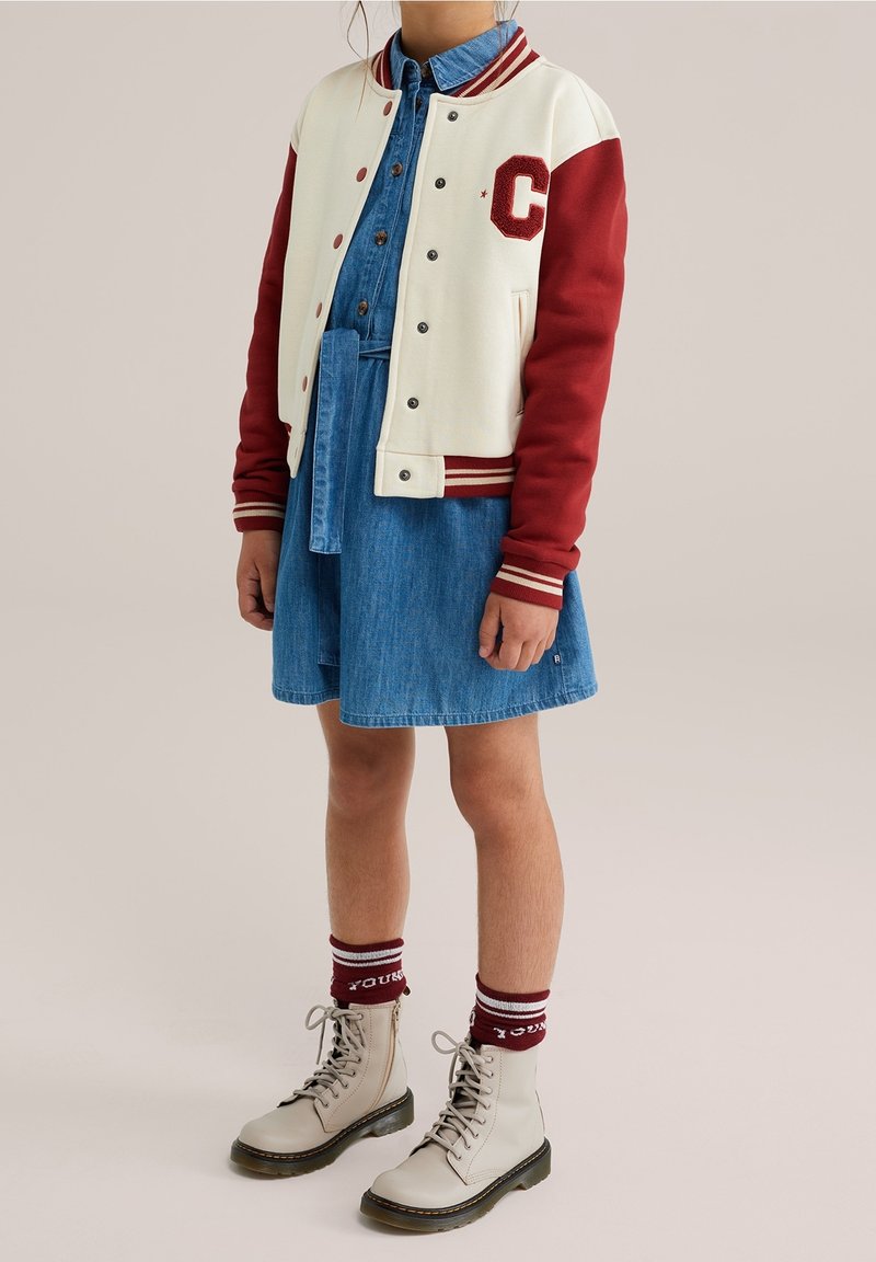 Dziecko ubrane w niebieską sukienkę dżinsową, kremowo-czerwoną kurtkę varsity z literą "C", bordowe skarpetki oraz beżowe sznurowane buty na jednolitym tle.