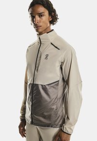 Jeune homme portant une veste légère zippée beige et grise à manches longues, se tenant devant un fond uni.