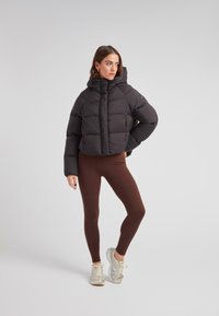 Czarna krótka kurtka puchowa z kapturem, z pikowanym wzorem i logo na rękawie, zestawiona z brązowymi legginsami i jasnymi sneakersami.