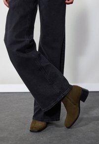 Botas de tobillo de ante marrón con puntera cuadrada y tacón cuadrado, combinadas con vaqueros de pierna ancha oscuros, que presentan un detalle de cremallera lateral.