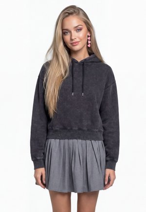 Jonge vrouw met lang blond haar, een donker hoodie, een grijze geplooide rok en roze oorbellen in hartvorm, staand tegen een witte achtergrond.