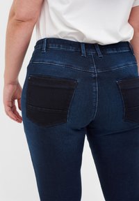 Jeans de mezclilla azul oscuro con bolsillos traseros negros en contraste. Presentan una cinturilla estándar y una forma ajustada, mostrando una textura suave.