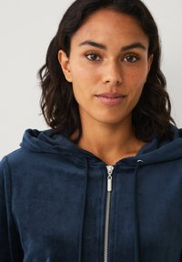 Sudadera con capucha azul marino con cremallera frontal, hecha de un tejido suave y texturizado. Presenta cordones y un detalle de cremallera plateada.