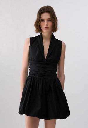 Donna con capelli corti e ondulati che indossa un vestito nero senza maniche con scollatura a V profonda e vita arricciata, in piedi davanti a uno sfondo grigio.