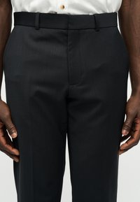 Κοντινή λήψη μαύρου επίσημου παντελονιού φορεμένου με λευκό πουκάμισο βαλμένο μέσα, που δείχνει θηλιές ζώνης και μπροστινές ραφές σε έναν όρθιο άνθρωπο.