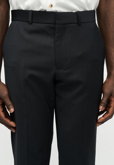 Gros plan sur un pantalon noir de cérémonie porté avec une chemise blanche rentrée, montrant les passants de ceinture et les coutures avant sur une personne debout.