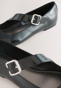 Schwarze Lack-Mary-Jane-Ballerinas mit silbernen Schnallenriemen und abgerundeten Spitzen auf hellem Hintergrund.