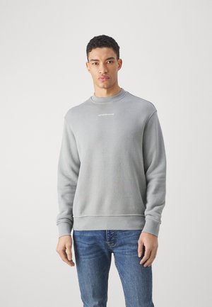 Felpa grigia in cotone con colletto rotondo, polsini e orlo a coste, caratterizzata da una discreta ricamo del logo sul petto. Indossata con jeans blu.