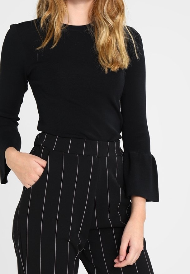 Czarna bluzka z długim rękawem i rozszerzanymi mankietami w połączeniu z czarnymi spodniami o wysokim stanie, ozdobionymi pionowymi białymi paskami i gładką teksturą.