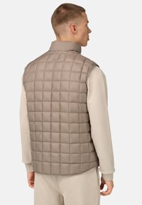 Vadderad väst i ljusbeige med quiltat mönster, hög krage och en slät yta. Bärs över en långärmad topp, vilket visar en minimalistisk stil.