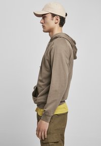Felpa marrone con tasca frontale e cordino, indossata sopra una maglietta gialla. Cappellino beige, pantaloni cargo verde oliva, visti di profilo.