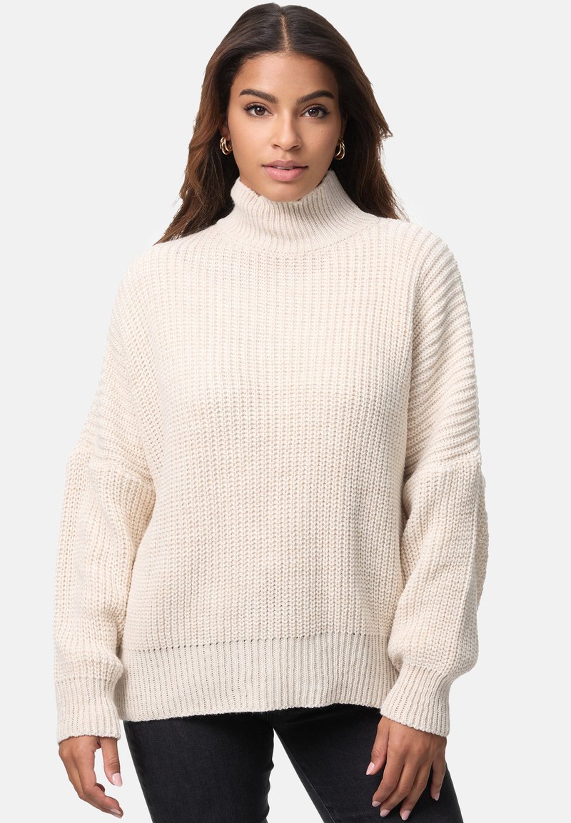 Kremfarvet strikket sweater med høj rullekrave, løse ærmer, ribstrikkede manchetter og et tekstureret mønster, kombineret med mørke jeans.