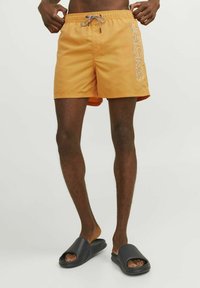 Bañador naranja hecho de material ligero, con cintura elástica ajustable y estampado de logo en el lateral. Chanclas negras completan el look.