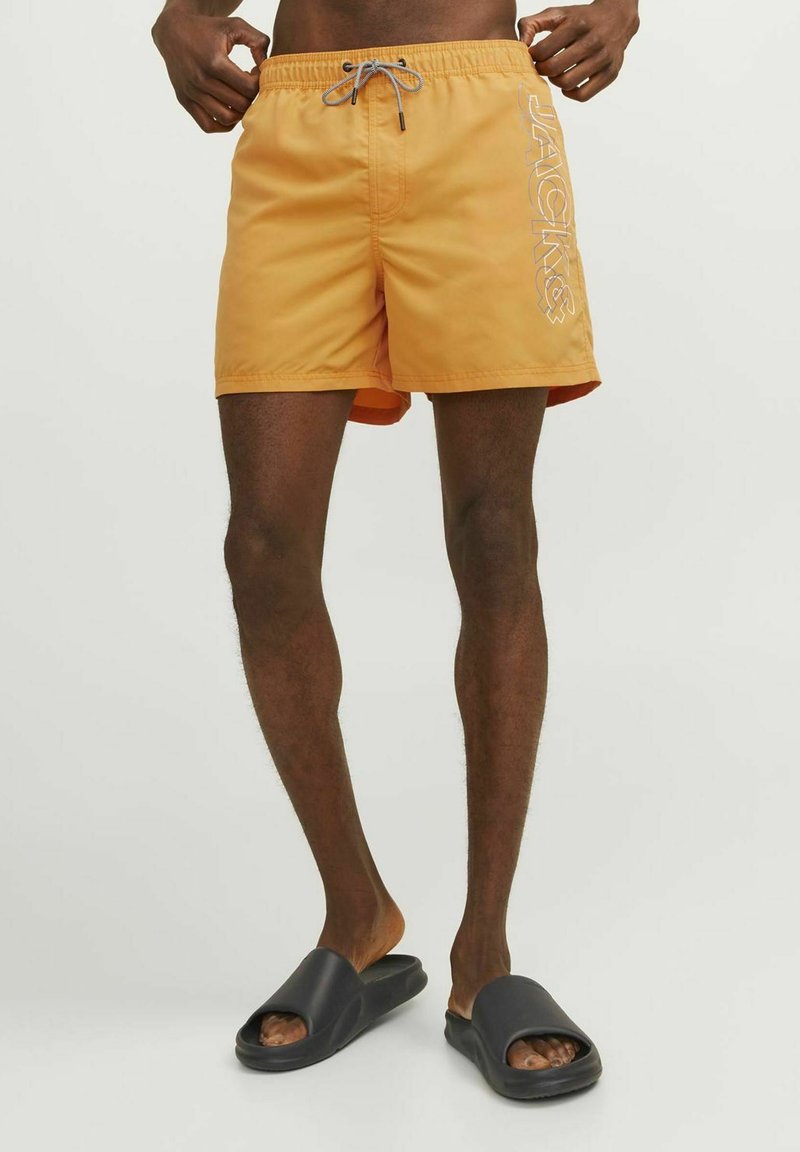 Bañador naranja hecho de material ligero, con cintura elástica ajustable y estampado de logo en el lateral. Chanclas negras completan el look.