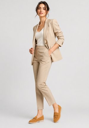 Frau steht und trägt einen beigen Blazer und Hose mit weißem Top und braunen Loafers vor einem einfarbigen hellen Hintergrund.
