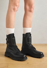 Botas de cuero negro hasta el tobillo con cordones frontales y suela de goma gruesa. Presentan acentos de tachuelas y correas con hebillas ajustables. Acabado texturizado.