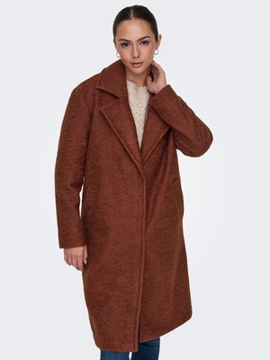 Femme portant un long manteau marron texturé sur un pull léger en maille, avec des boucles d'oreilles créoles et sa main touchant son cou.