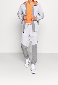 Survêtement léger gris à motif quadrillé, composé d'une veste à capuche orange et d'un pantalon ajusté. Orné de détails gris et blancs.