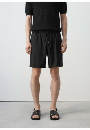 Hombre con camiseta de punto negra de manga corta, pantalones cortos negros con cordón y sandalias negras abiertas, de pie contra una pared gris lisa.