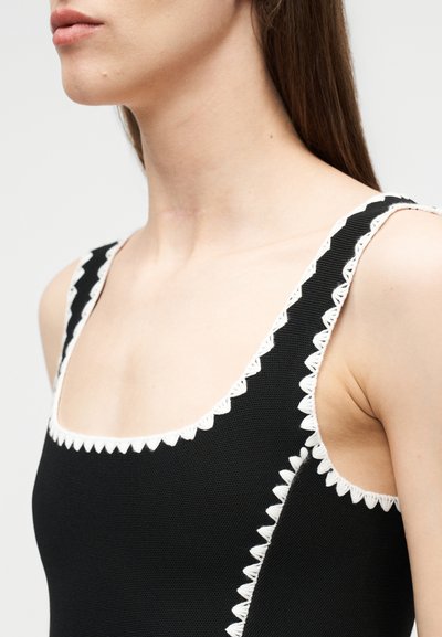 Jeune femme portant un haut noir sans manches avec une bordure blanche en forme de festons le long du décolleté et des bretelles, vue de la tête aux épaules.