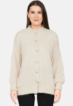 Vrouw met recht bruin haar, gekleed in een beige grofgebreide gebuttonde cardigan en zwarte broek, kijkend naar voren tegen een witte achtergrond.