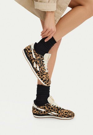 Persona con zapatillas Reebok con estampado de leopardo y calcetines negros, ajustándose un zapato con las manos, pantalones cortos beige visibles sobre fondo claro.