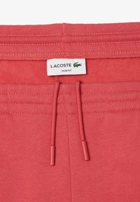 Pantalons de survêtement slim fit corail avec une texture douce, taille élastique avec cordon de serrage et embouts revêtus, et étiquette blanche Lacoste.