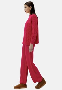 Różowy sweter dzianinowy z wzorem w warkocze, połączony z dopasowanymi luźnymi spodniami i brązowymi botkami za kostkę. Gładka tekstura i swobodny fason.
