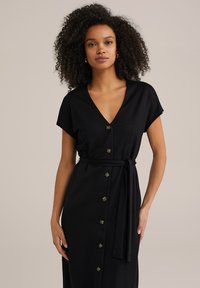 Abito nero a camicia con maniche corte, caratterizzato da una scollatura a V e una cintura in vita. Tessuto liscio e grandi bottoni rotondi lungo la parte anteriore.