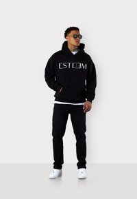 Haut à capuche noir avec logo "ESTEEM" blanc, poche avant, porté par-dessus une chemise blanche, associé à un jean noir déchiré et des baskets blanches.