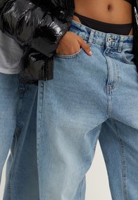 Svart puffjacka över en figursydd grå skjorta, tillsammans med ljusblå denimjeans i avslappnad passform och en klassisk femfickdesign.