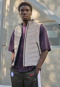 Beige padded vest with a high collar and black zipper, worn over a dark purple t-shirt and black pants with Nike and PSG logos.