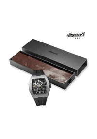 Zwarte analoge horloge met een transparante wijzerplaat en zilveren accenten, gemonteerd op een zwarte rubberen strap, weergegeven in een zwarte doos met bruine voering.