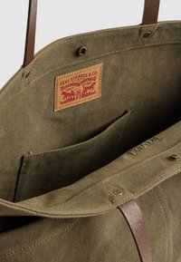 Olivgrüne Canvas-Umhängetasche von Levi Strauss mit braunen Ledergriffen, Innenfach und genähtem Logopatch innen.
