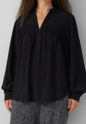 Frau trägt eine locker sitzende schwarze Bluse mit gerafftem Einsatz und langen, mit Manschetten versehenen Ärmeln, kombiniert mit grauen, strukturierten Hosen.