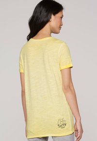 Kurzärmliges gelbes T-Shirt aus strukturiertem Stoff, mit kontrastierenden Saumabschlüssen an den Ärmeln und einem aufgedruckten Design nahe dem Saum, das "Soccy Stores" detailiert.