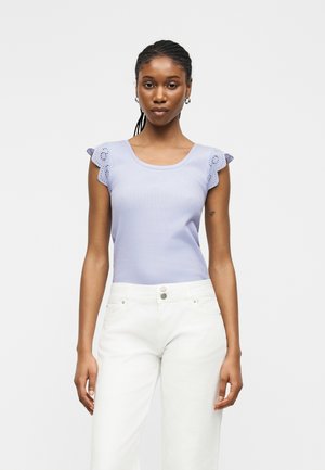 Γυναίκα με πλεξούδες, φορόντας ένα αμάνικο ανοιχτό μωβ τοπ με βολάν στα μανίκια και λευκά ψηλόμεσα παντελόνια, με απλό φόντο.