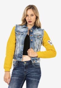Gilet en denim avec tissu déchiré, de couleur bleu clair, et manches longues jaunes. Présente des boutons en métal et un patch logo sur la poitrine.