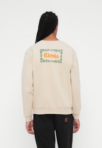 Beżowy sweter z okrągłym dekoltem, z kwiecistym wzorem i napisem "Rivvia" w pomarańczowym kolorze na plecach. Czarne spodnie z pomarańczowym akcentem.