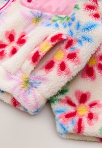 Manche de veste douce et moelleuse avec des motifs floraux colorés en rouge, rose, jaune, bleu et vert sur fond blanc, montrant une ouverture de poche.