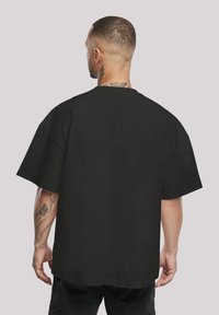 Melns oversize T-krekls ar apļa kakla izgriezumu, īsām piedurknēm un kokvilnas materiālu. Gluda tekstūra ar brīvu piegriezumu, skatīts no aizmugures.