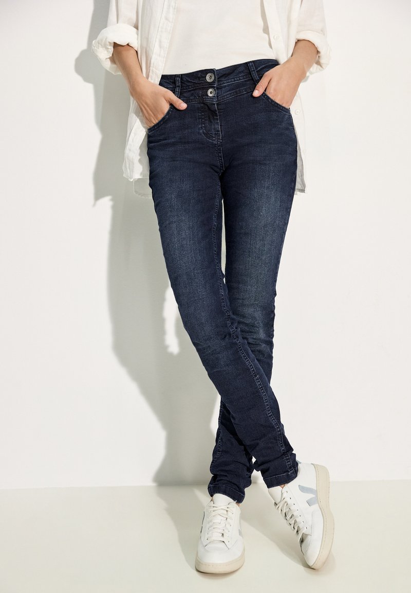 Jean en denim bleu foncé avec une coupe skinny, dotés d'une fermeture à double bouton et de poches avant, associés à des baskets blanches.