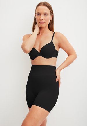 Vrouw draagt zwarte beha en zwart corrigerend shorts met hoge taille, poseert met één hand bij haar nek en de andere op haar heup tegen een witte achtergrond.