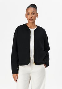 Schwarze Zip-Jacke aus weichem Material mit Rundhalsausschnitt, zwei Fronttaschen und goldenen Reißverschlüssen. Über einem weißen Oberteil getragen.