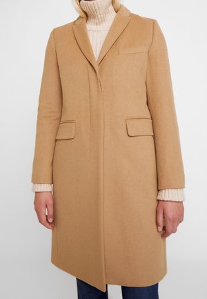 Vrouw met een knie-lange camel kleurige jas over een crèmekleurige coltrui, staand met de handen ontspannen langs haar zij.