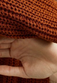 Rust-colored knitted fabric with an open weave texture, showcasing a soft hand resting beneath it, emphasizing the material's drape.