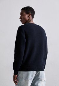 Navy gestrickter Pullover mit schräg geripptem Muster und Rundhalsausschnitt. Kombiniert mit hellen, gewaschenen Jeans, die ein minimalistisches Design aufweisen.