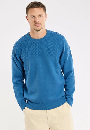 Homme aux cheveux courts portant un pull bleu texturé avec des boutons sur les épaules et un pantalon beige, debout les mains dans les poches.